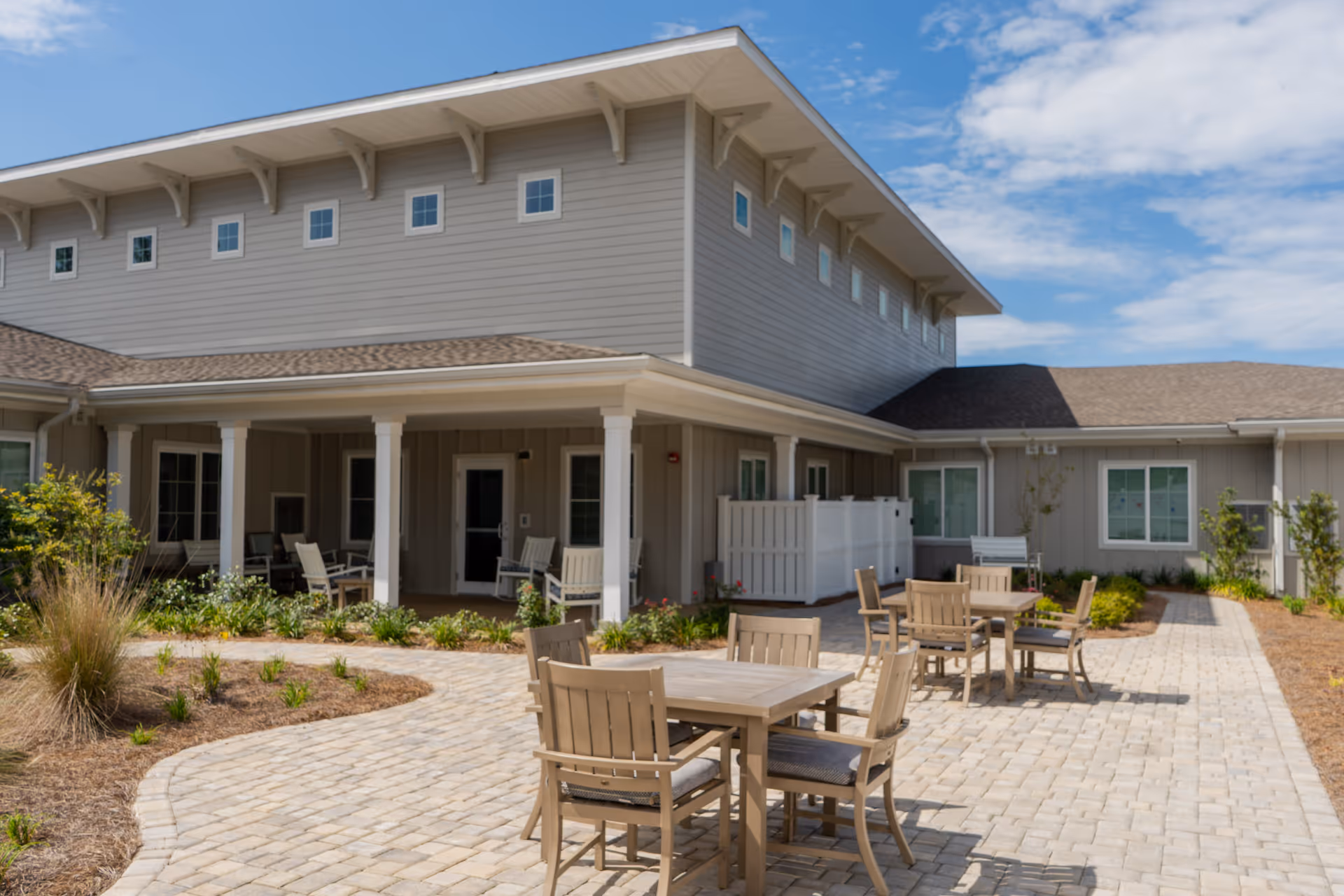 Outdoor patio area at The Landing of Panama City Beach featuring several wooden tables and chairs on a paved surface, surrounded by landscaped plants and shrubs, with a two-story building in the background under a partly cloudy sky.