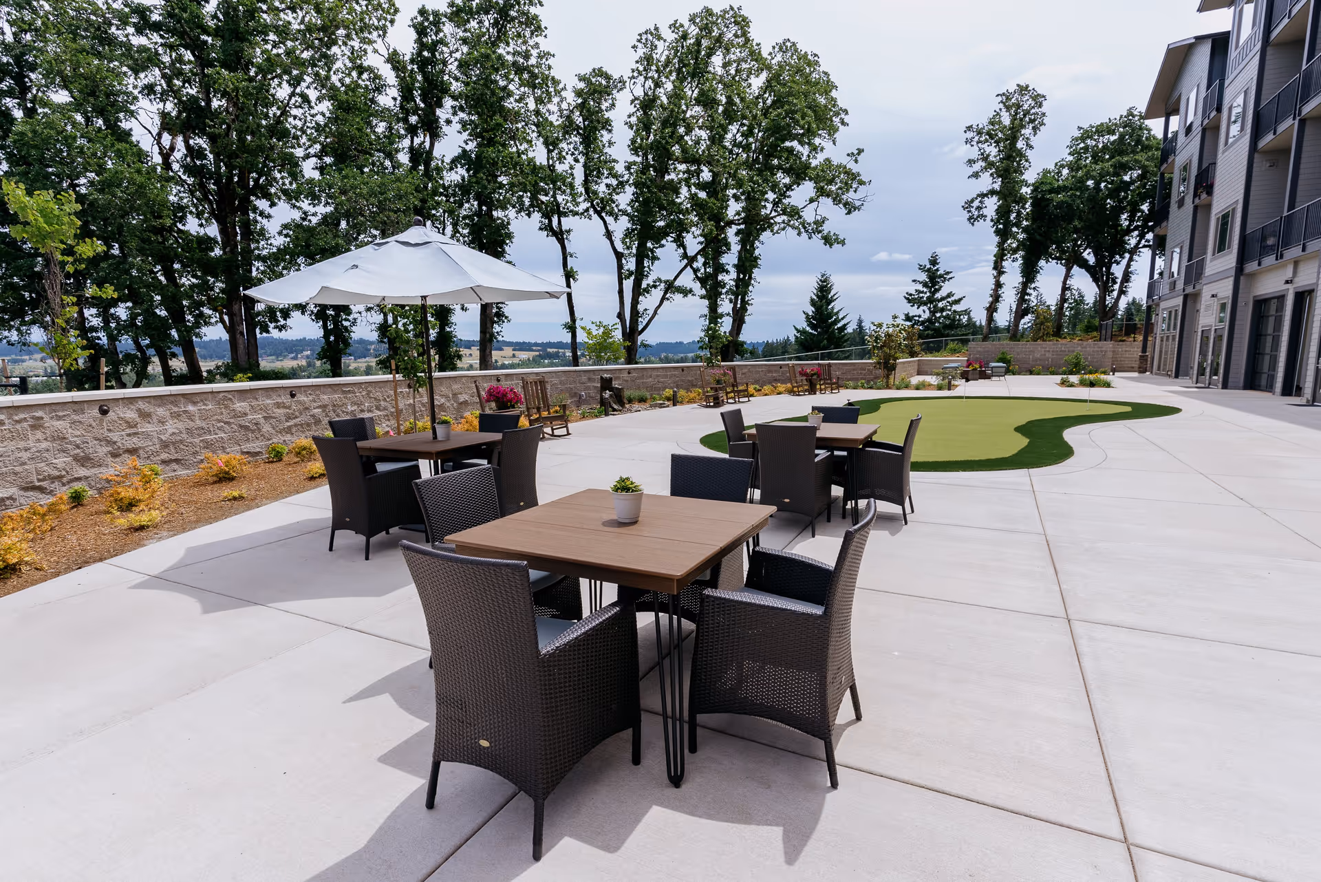 Outdoor patio area at Boone Ridge Retirement Community featuring several tables with wicker chairs, one table shaded by a large white umbrella, a small putting green, and surrounded by trees and landscaping.