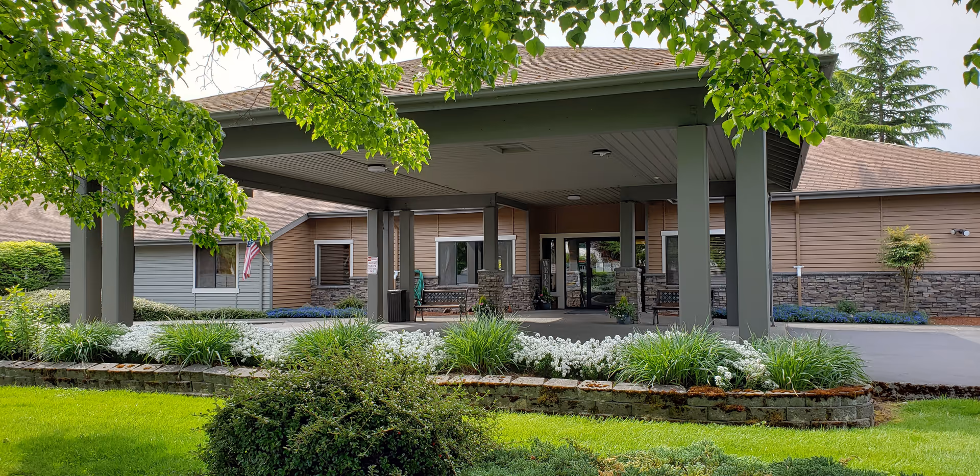 Entrance of Village Concepts of Marysville - Grandview Village showing a covered drop-off area with pillars, surrounded by green grass, bushes, and white flowers. The building has a brown and gray exterior with stone accents and windows, and an American flag is visible on the left side.