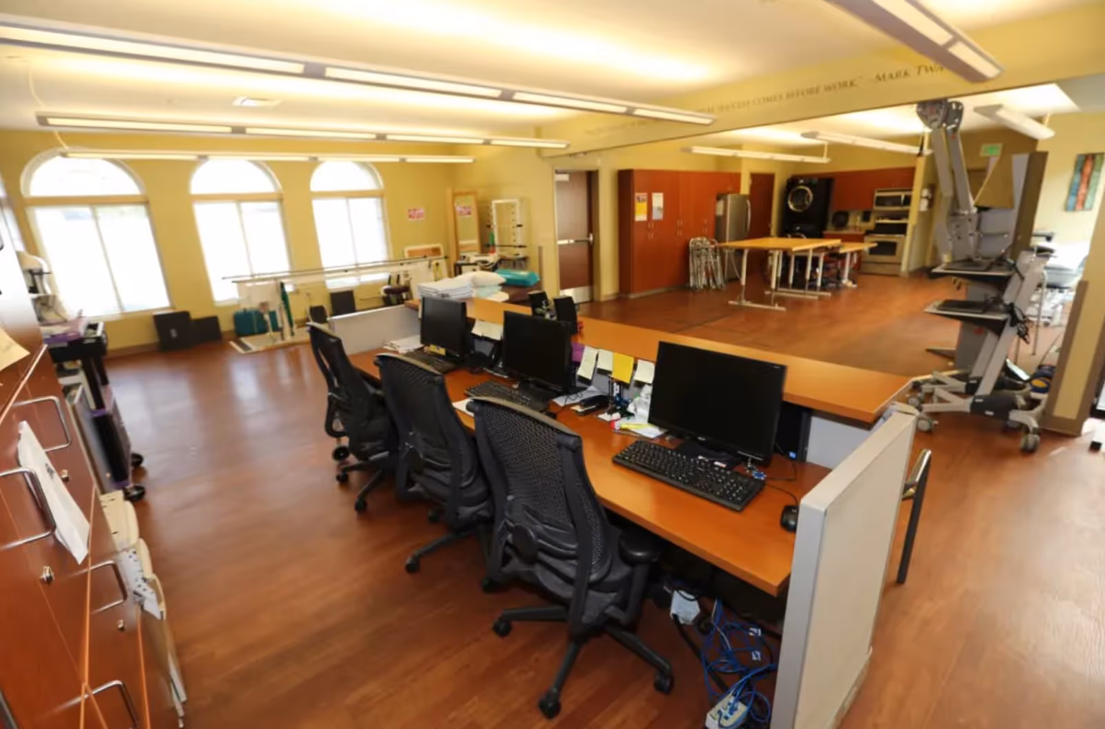 Interior view of a senior living facility office area with a row of desks and computer workstations in the foreground. The room has large arched windows letting in natural light, wooden flooring, and various medical and rehabilitation equipment in the background.