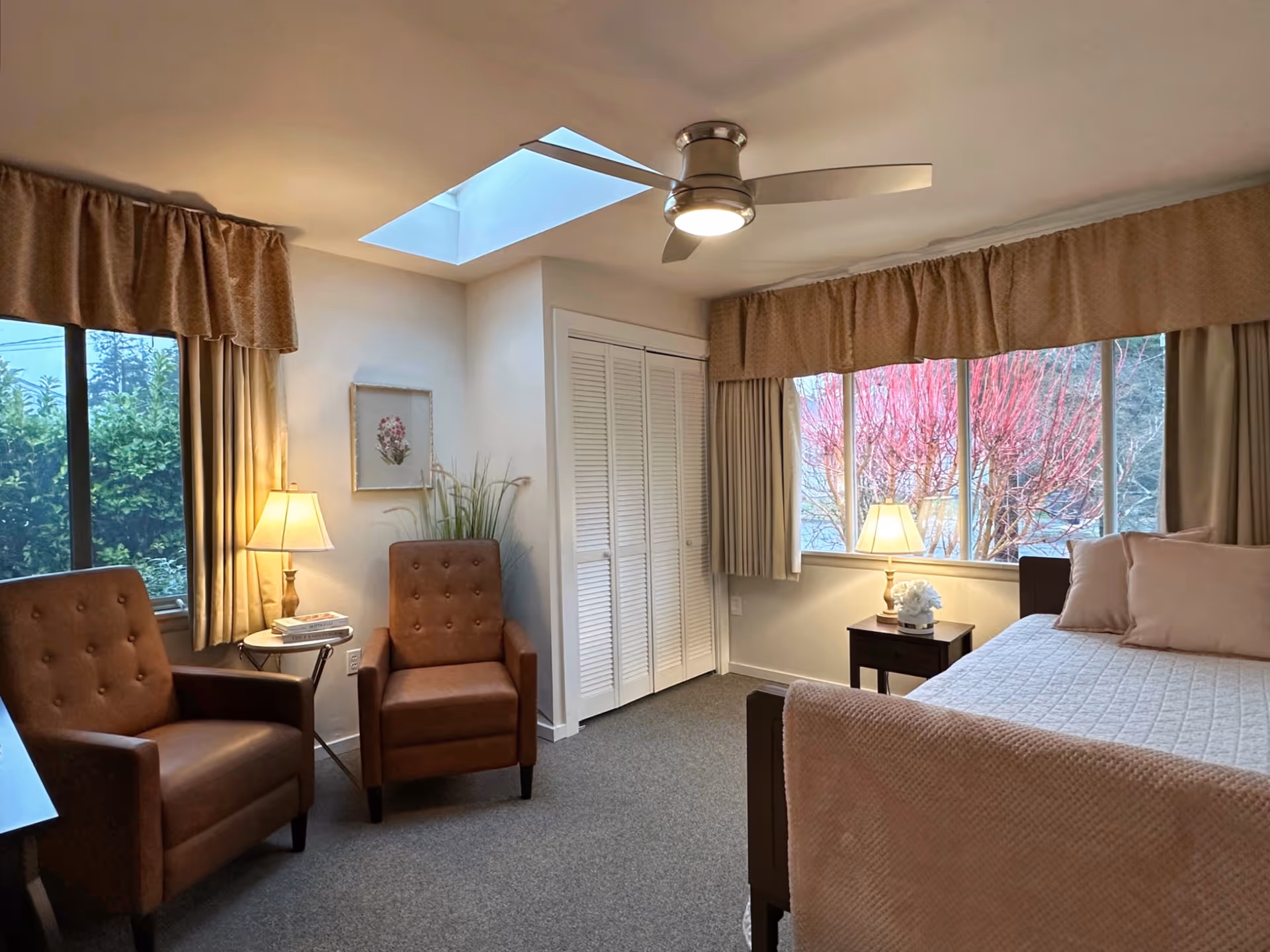A bright bedroom with two brown armchairs, a bed with light bedding, bedside tables and lamps, large windows with curtains and a ceiling fan.