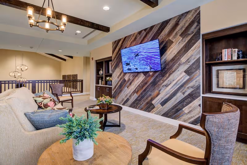A cozy living room area with a beige sofa adorned with colorful pillows, a wooden round coffee table with a potted plant, and a wooden accent wall with a mounted flat-screen TV displaying an image of ducks on water. The room features exposed wooden ceiling beams, built-in dark wood shelving with decorative items, and warm lighting fixtures including a chandelier and pendant lights.