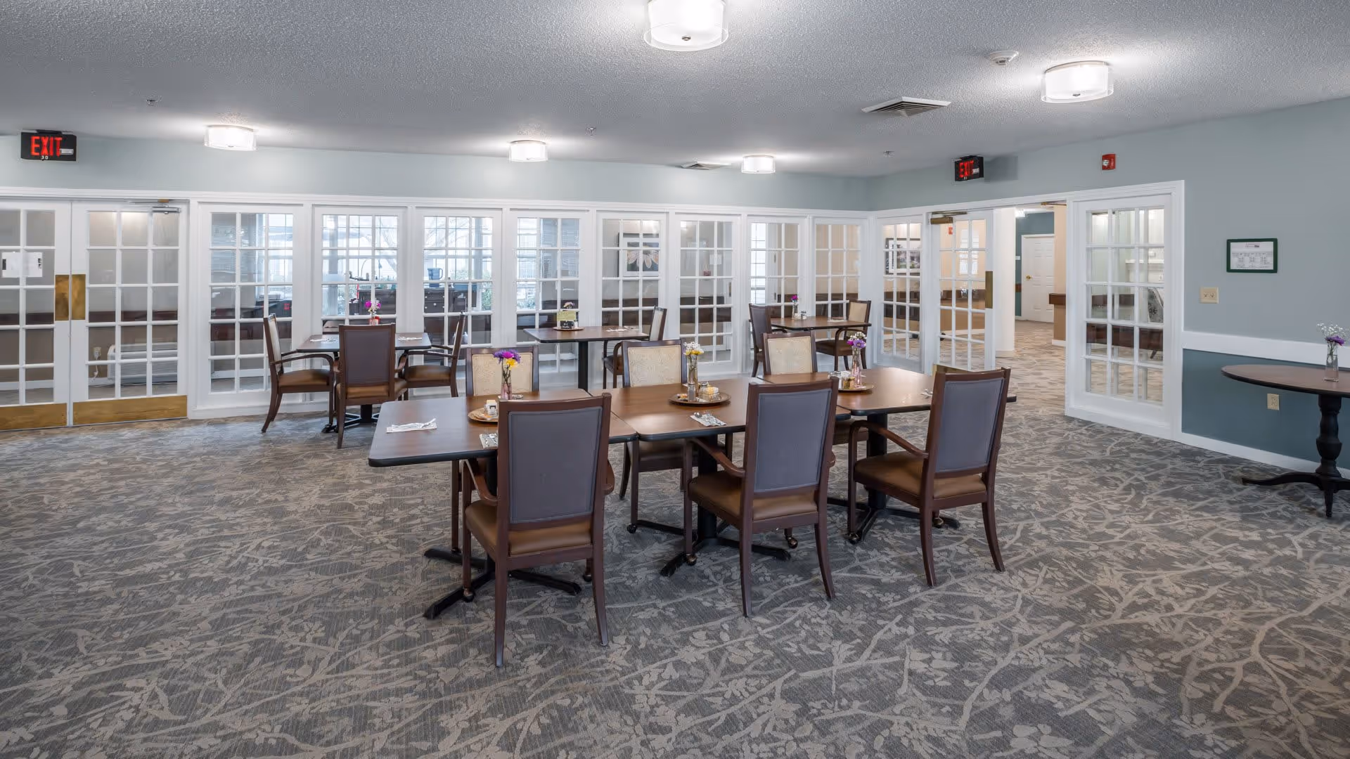 A spacious dining room with multiple wooden tables and chairs arranged neatly. Each table has small flower vases as centerpieces. The room has light blue walls, patterned carpet flooring, and large glass-paneled doors and windows along the walls. Ceiling lights provide bright illumination, and exit signs are visible above the doors.