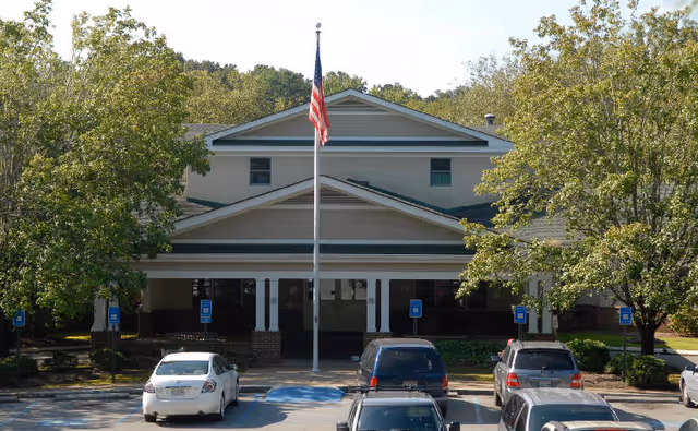 Front exterior view of a two-story building with a covered entrance, an American flag on a flagpole in front, surrounded by trees and a parking lot with several cars parked.
