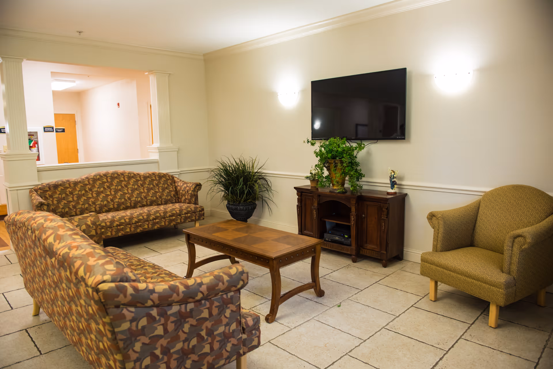 A cozy living room area with two patterned sofas, a single armchair, a wooden coffee table, a wooden TV stand with a potted plant and decorative items, and a flat-screen TV mounted on the wall. The room has tiled flooring and soft lighting from wall sconces.