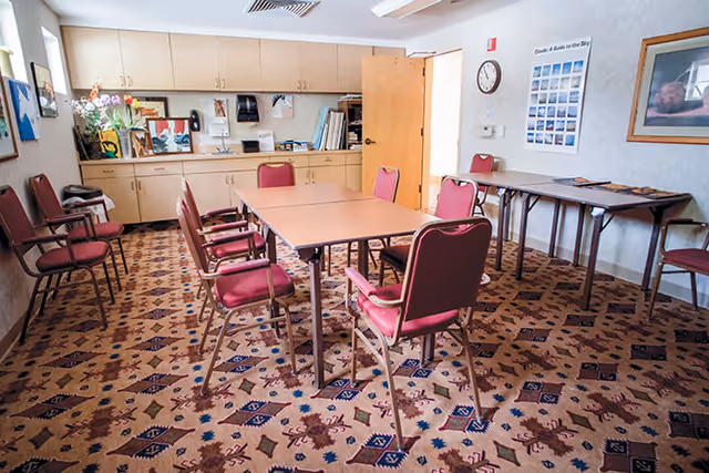 A meeting or activity room with a rectangular table surrounded by red cushioned chairs on a patterned carpet. The room has beige cabinets along one wall with various items on the countertop, including flowers and framed pictures. There is a clock and a poster on the wall, and an open door leading to another room.