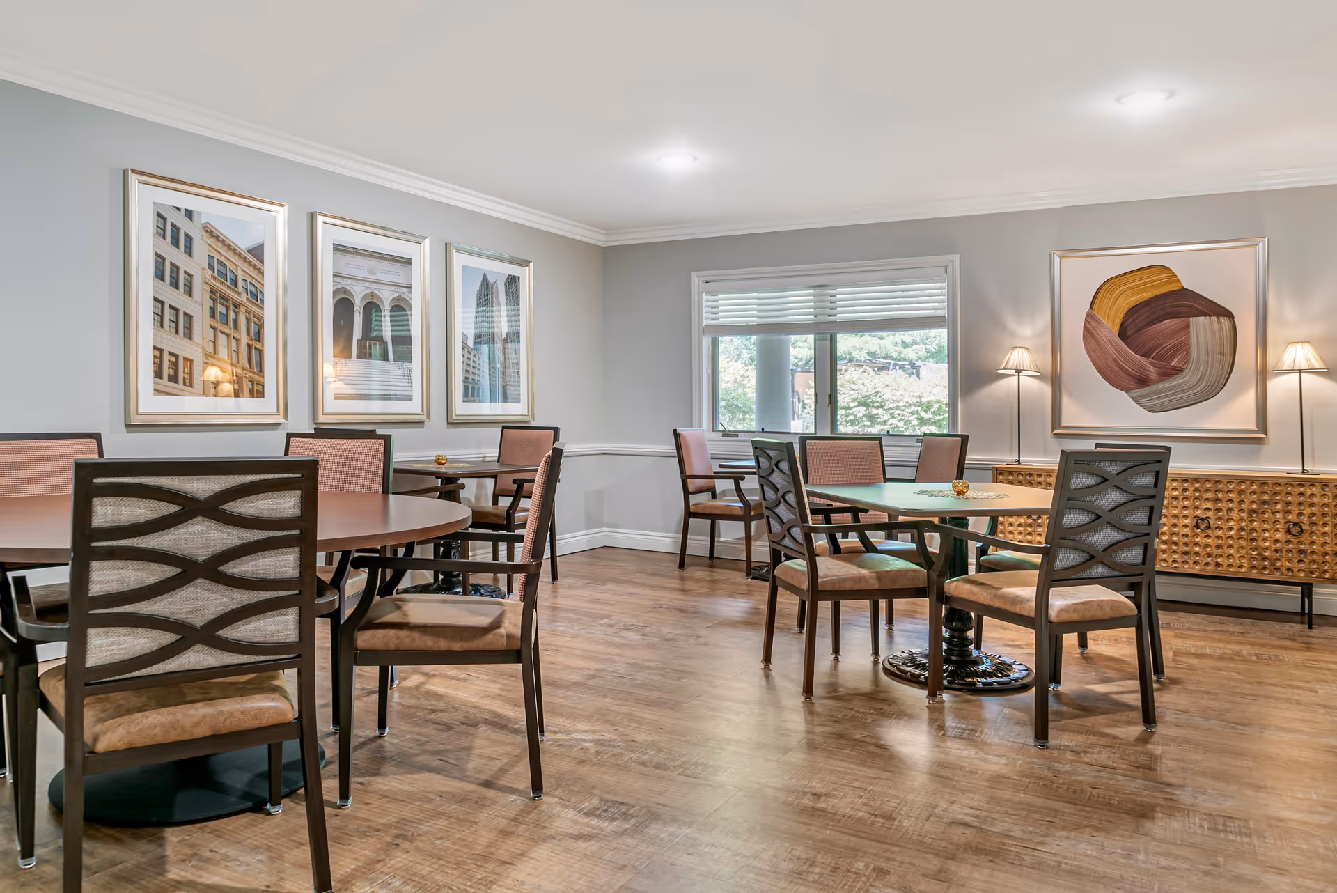 A well-lit dining room with wooden floors, round tables, and cushioned chairs. The walls are decorated with framed artwork and there is a window letting in natural light. Two floor lamps flank a wooden sideboard with a decorative front panel.