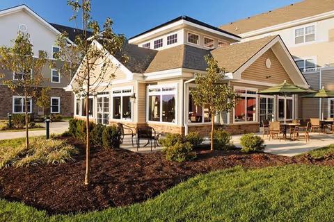 Exterior view of a senior living facility building with beige siding, large windows, and a patio area with tables and chairs. The foreground features landscaped garden beds with small trees and shrubs, and a well-maintained green lawn under a clear blue sky.