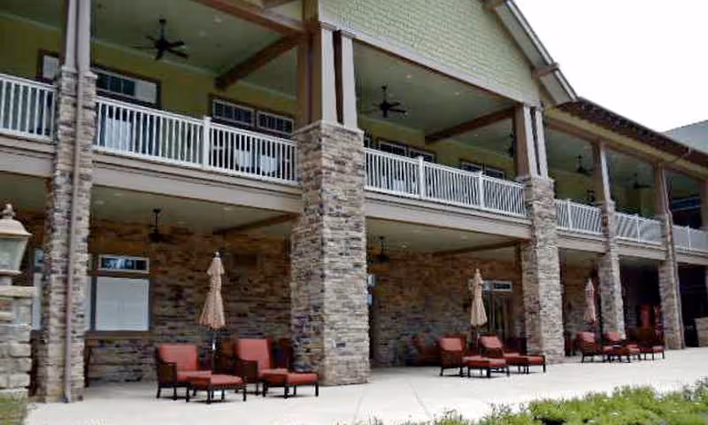 Outdoor patio area of a building with stone pillars and a covered balcony above. Several sets of cushioned chairs and umbrellas are arranged along the patio.