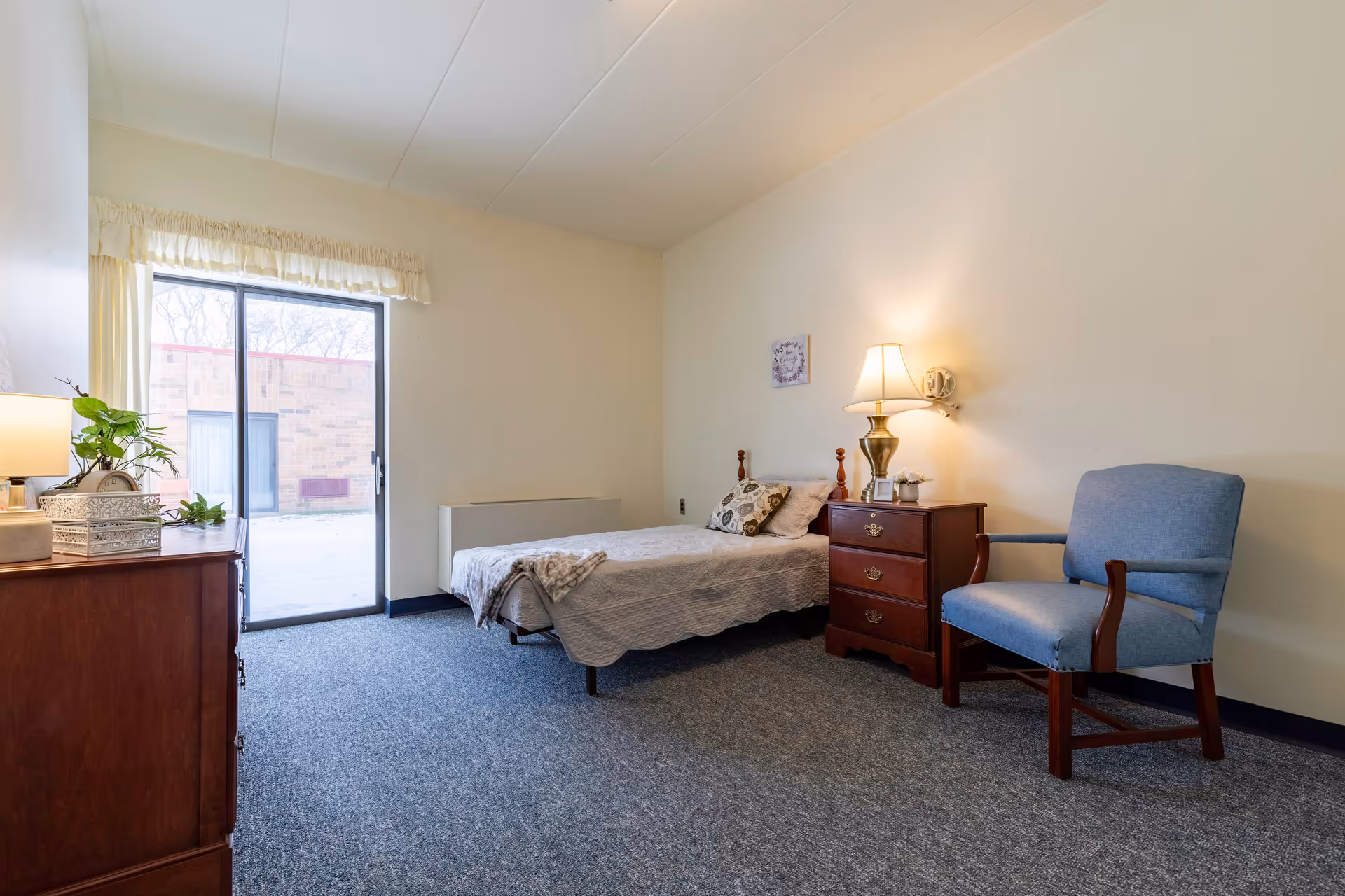 A simple bedroom in a senior living facility with a single bed covered in a light gray quilt, a wooden nightstand with a lamp and small decor items, a blue upholstered armchair, a wooden dresser with a lamp and plants, and a sliding glass door leading outside.