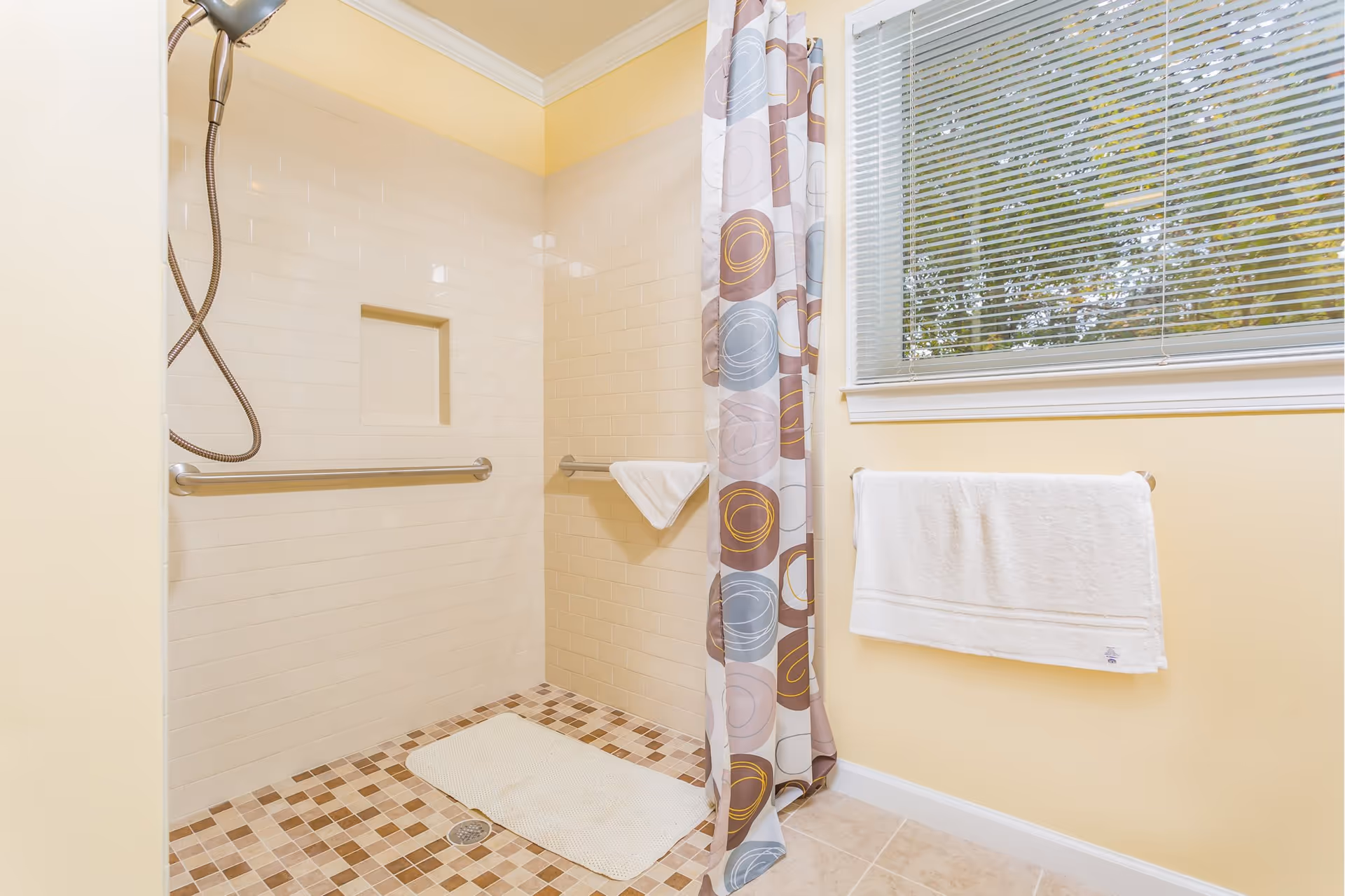 A bright bathroom shower area with beige tiled walls and a mosaic tile floor. The shower has a handheld showerhead, two horizontal grab bars, and a built-in niche for toiletries. A colorful shower curtain with circular patterns is partially drawn to the side. A white towel hangs on a towel rack beneath a window with closed blinds.