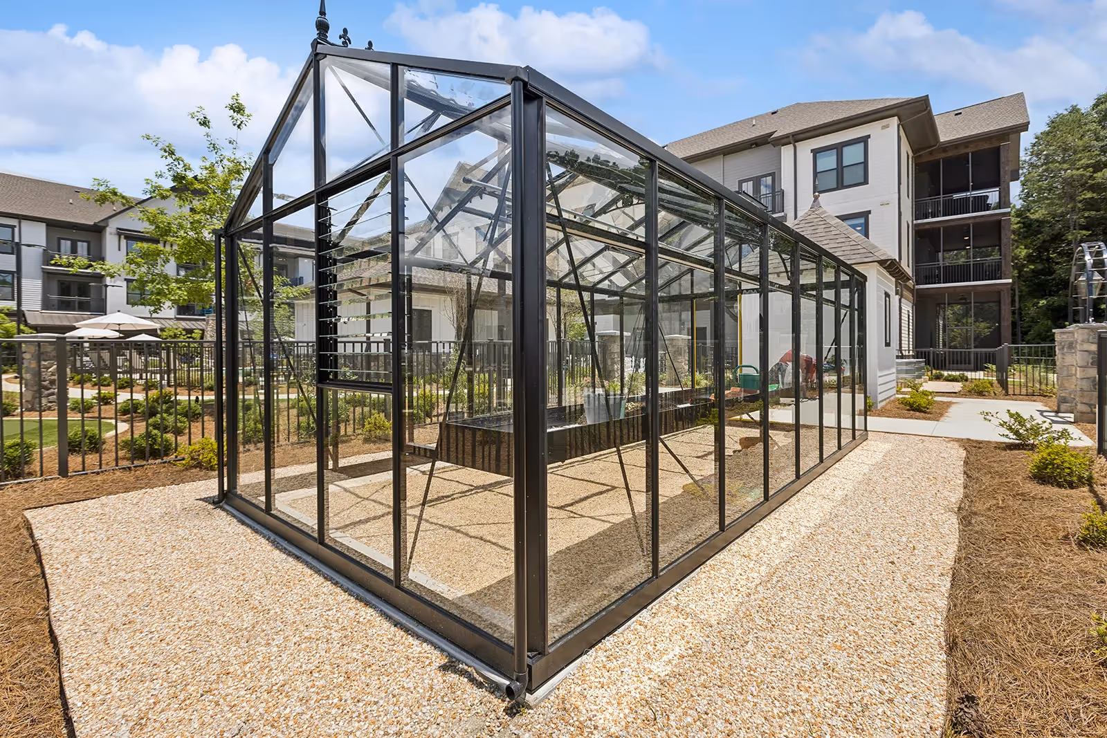A modern glass greenhouse structure situated outdoors on a gravel path, surrounded by landscaped garden beds and a multi-story residential building in the background under a blue sky with some clouds.
