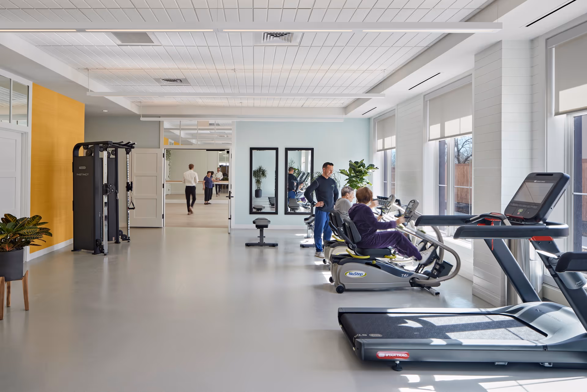 Bright fitness room with treadmills, recumbent bikes, a weight machine, and people exercising near large windows.