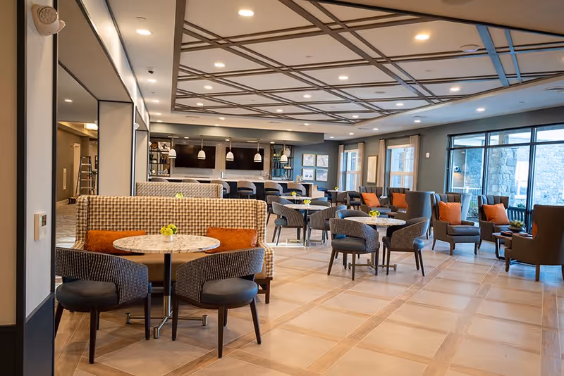 Spacious, well-lit communal dining and lounge area with multiple round tables, upholstered chairs and booths, and large windows.