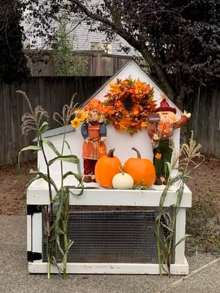Autumn display with three pumpkins, two scarecrow figures, and a fall wreath on a small white wooden box in front of a fence.