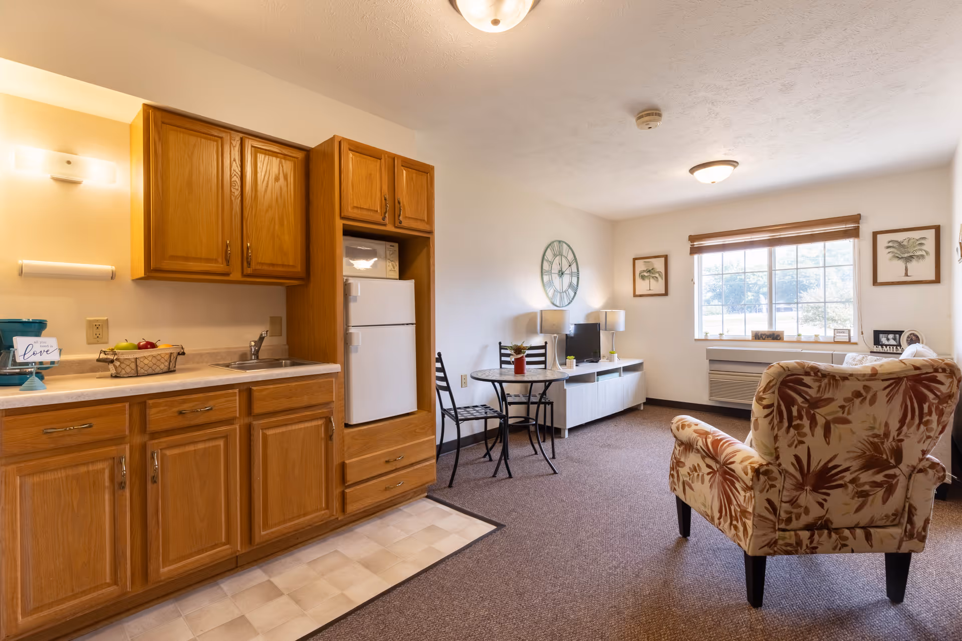 A cozy senior living apartment interior featuring a small kitchen area with wooden cabinets, a white refrigerator, and a sink. Adjacent to the kitchen is a small round dining table with two chairs. The living area includes a floral-patterned armchair, a white TV stand with a television, two table lamps, and wall decorations including a large clock and framed artwork. A window with blinds allows natural light into the room.