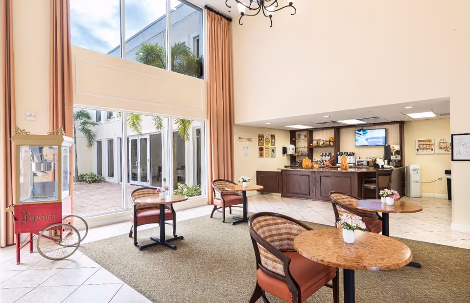 A bright and welcoming common area with large floor-to-ceiling windows letting in natural light. The room features several round tables with cushioned chairs, each table decorated with small flower arrangements. To the left, there is a vintage-style popcorn machine. In the background, there is a counter area with a coffee and beverage station, decorated with seasonal pumpkins and other items. The space has a high ceiling with a chandelier and light-colored walls and flooring.