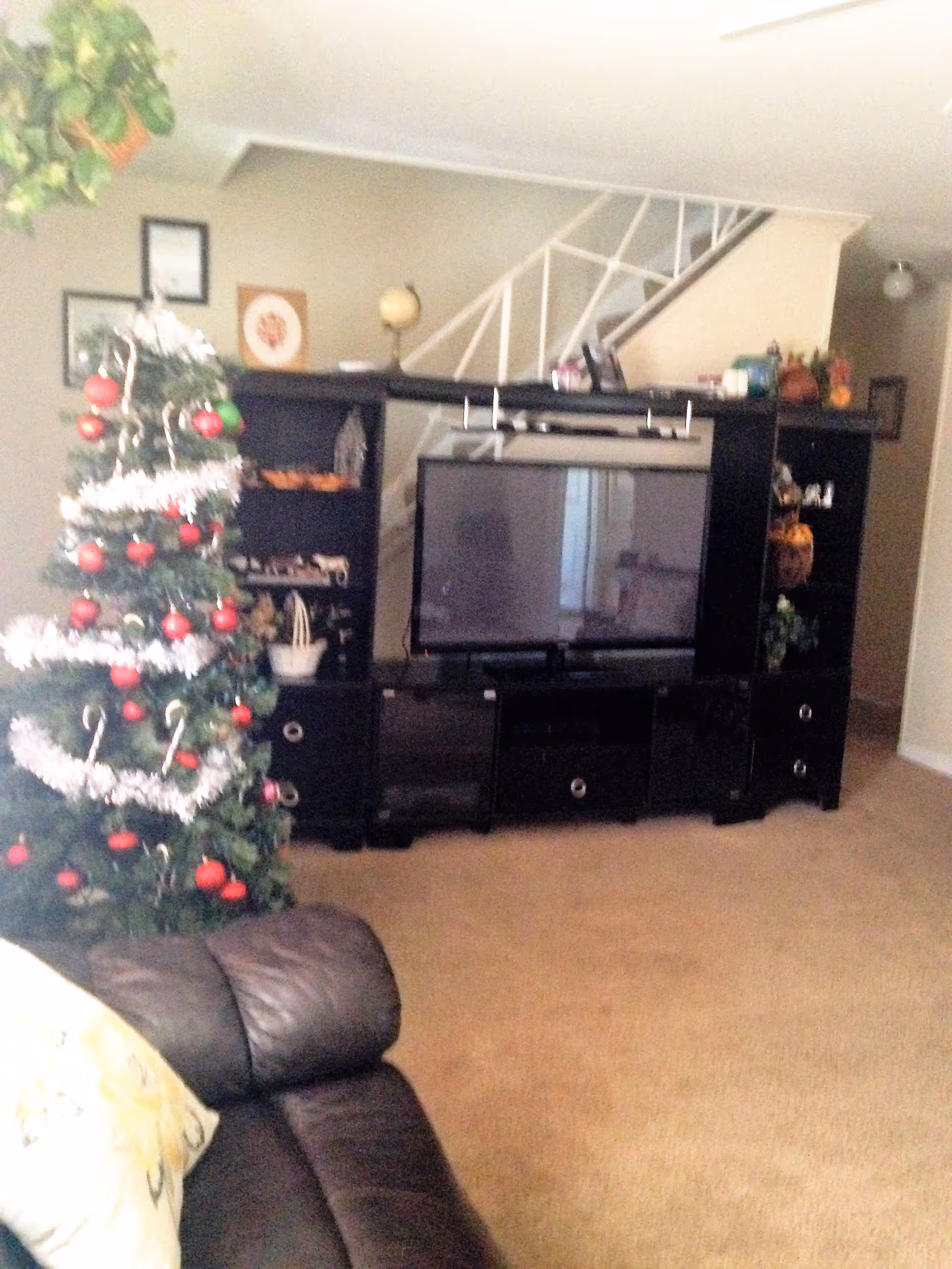Living room with a decorated Christmas tree on the left, a black entertainment center with a flat-screen TV in the center, and a dark leather couch with a floral pillow in the foreground. A staircase with white railing is visible in the background.
