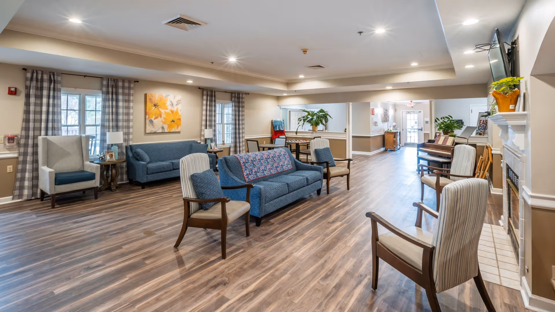 A spacious and well-lit common area in a senior living facility with wooden flooring, multiple seating options including sofas and armchairs, a fireplace with a TV mounted above it, potted plants, and large windows with checkered curtains allowing natural light to enter.