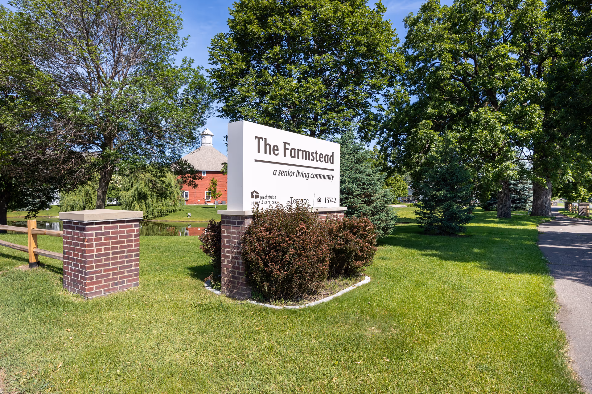 Outdoor view of The Farmstead senior living community sign surrounded by green grass, bushes, and trees with a red barn-style building in the background under a clear blue sky.