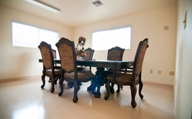 A dining room with a dark wooden dining table surrounded by six ornate upholstered chairs. The room has light-colored walls and two large windows letting in natural light. A floral centerpiece is placed on the table.
