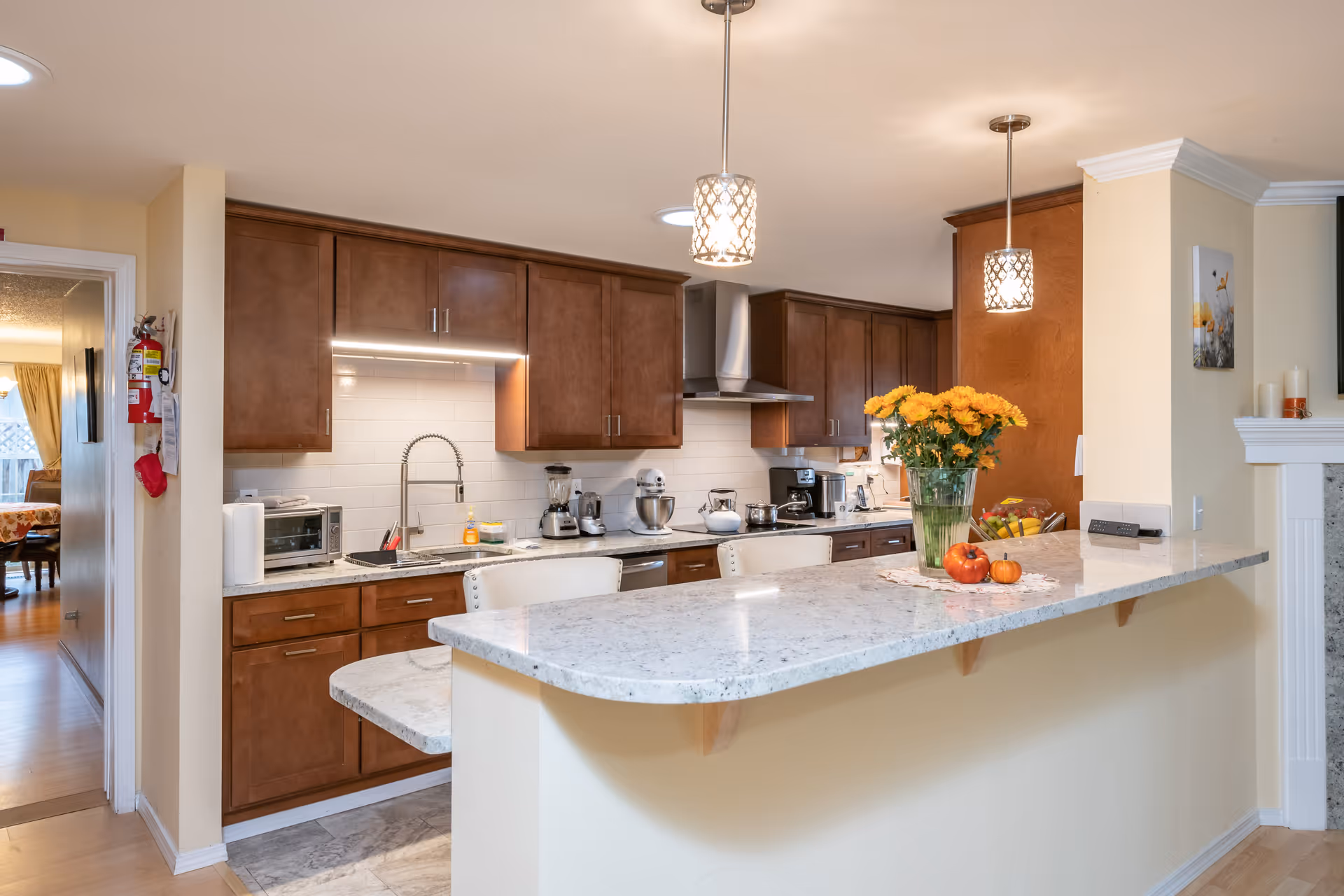A modern kitchen with brown wooden cabinets, a white marble countertop island with two white chairs, and stainless steel appliances including a toaster oven, blender, mixer, and coffee maker. There are two pendant lights hanging above the island, a vase with yellow flowers, and small decorative pumpkins on the countertop. The kitchen opens into a hallway leading to another room with a dining table and chairs.