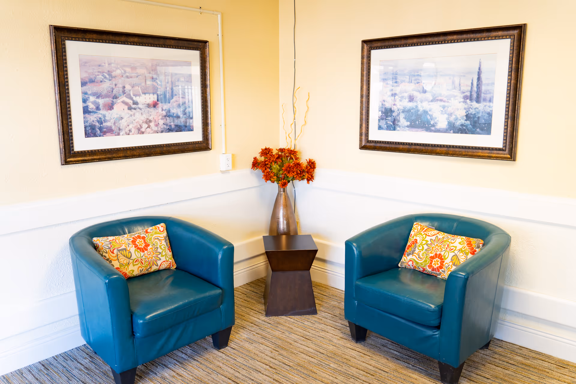 Two teal armchairs with colorful floral cushions are placed in a corner of a room with beige walls. Between the chairs is a small dark wooden side table holding a tall vase with orange flowers and decorative branches. Above each chair hangs a framed landscape painting.