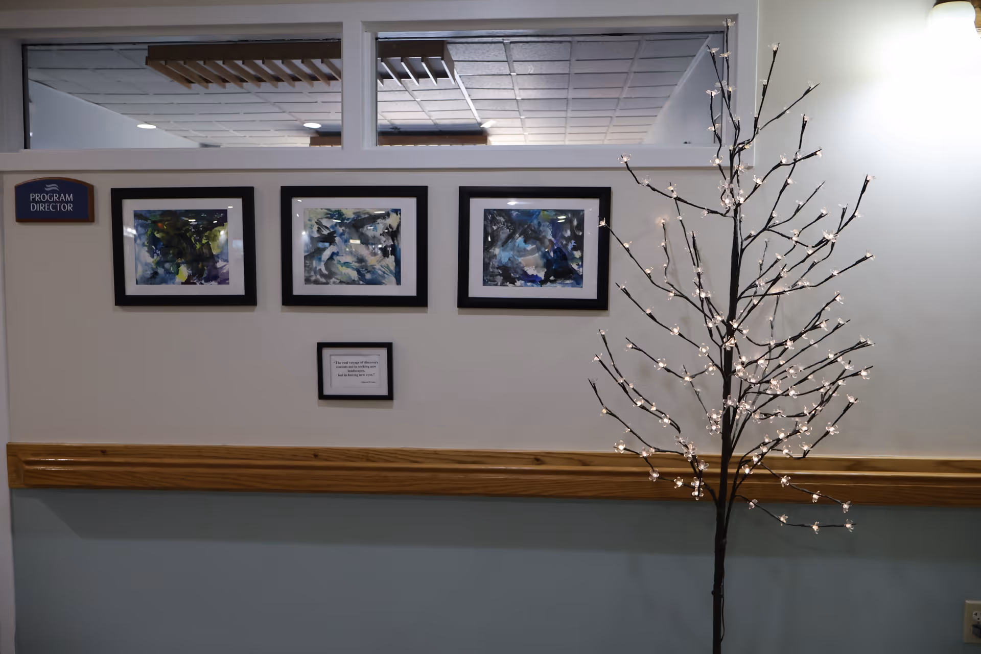 A hallway wall in a senior living facility with three abstract paintings framed in black, a small framed quote below them, a sign labeled 'PROGRAM DIRECTOR' on the left, and a decorative lighted tree with small white lights on the right.
