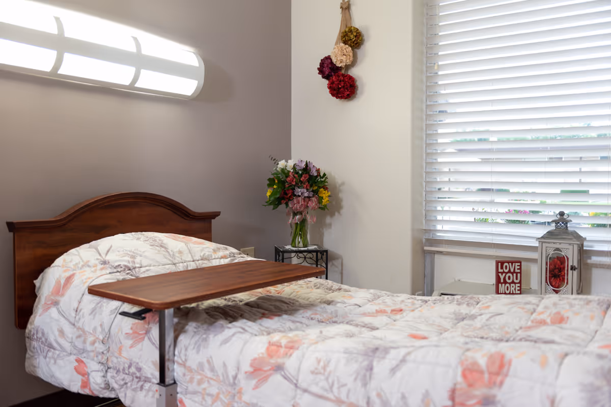 A single bed with a floral comforter and a wooden headboard in a room. A wooden overbed table is positioned over the bed. On the right side, there is a window with white blinds, a small table with a vase of colorful flowers, a decorative lantern, and a small sign that reads 'LOVE YOU MORE'. Above the bed, a modern wall light fixture is mounted, and a small hanging decoration with pom-poms is on the wall.