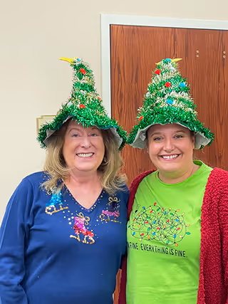 Two women smiling indoors wearing festive Christmas tree hats. One woman is wearing a blue sweater with holiday designs, and the other is wearing a green shirt with a holiday lights graphic and a red cardigan. A wooden door is visible in the background.
