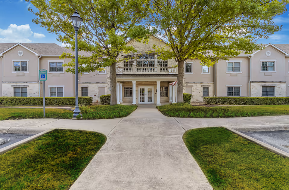 Front exterior view of Gemstone Senior Living at Castle Hills building with a paved walkway leading to the entrance, flanked by green grass and two large trees. The building has beige walls, multiple windows, and a covered porch with white columns.