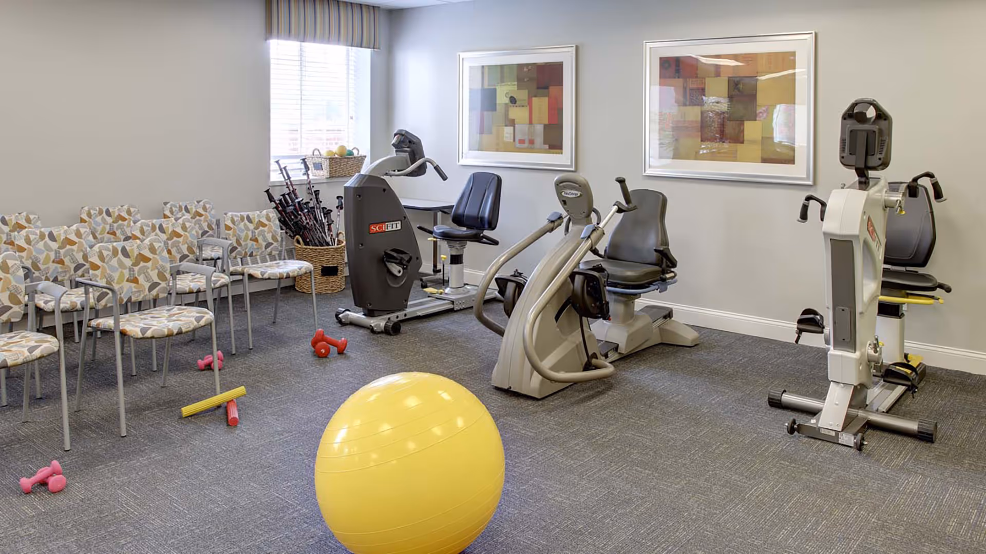 A fitness room with exercise equipment including stationary bikes and a rowing machine, a large yellow exercise ball, small pink dumbbells, and chairs with patterned cushions arranged along the wall. Two framed abstract artworks hang on the wall above the exercise machines, and a window with blinds lets in natural light.