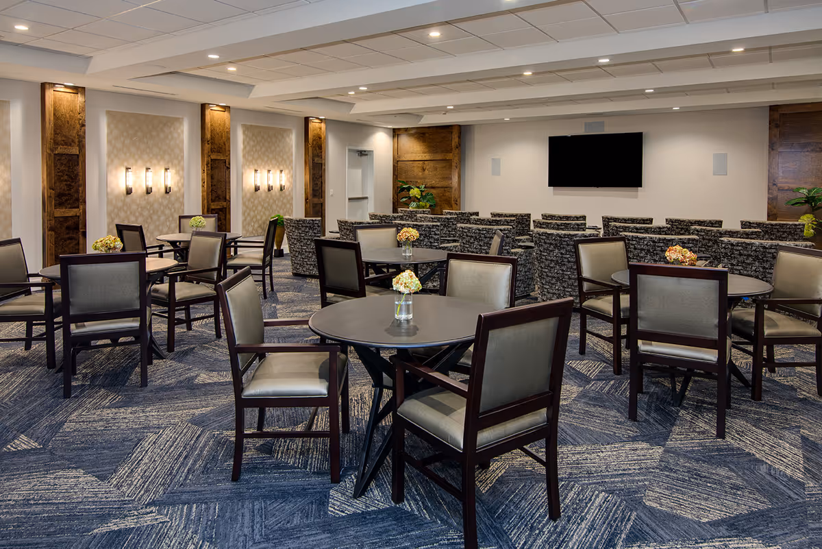 A spacious senior living common area with multiple round tables and chairs arranged for group seating. The room features patterned carpet flooring, decorative wall sconces, wooden panel accents, and a large flat-screen TV mounted on the far wall. Small floral centerpieces are placed on each table.