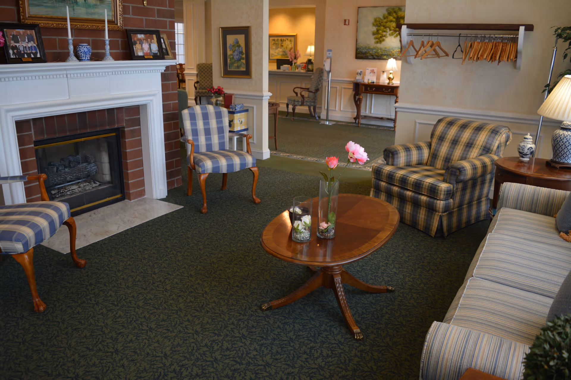 A cozy living room area with a green patterned carpet, a wooden coffee table with glass vases containing flowers, a plaid armchair, a striped sofa, and two wooden chairs with blue and white checkered cushions. There is a white fireplace with a brick surround, framed pictures and a blue vase on the mantel. The background shows a hallway with additional seating and framed artwork on the walls.