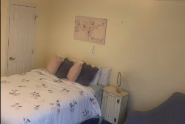 A cozy bedroom with a bed covered in a white floral comforter and several pillows in shades of pink and gray. There is a small white nightstand next to the bed with a decorative item on top. A door is visible on the left side of the room, and a piece of floral wall art hangs above the bed. The walls are painted a light yellow color.