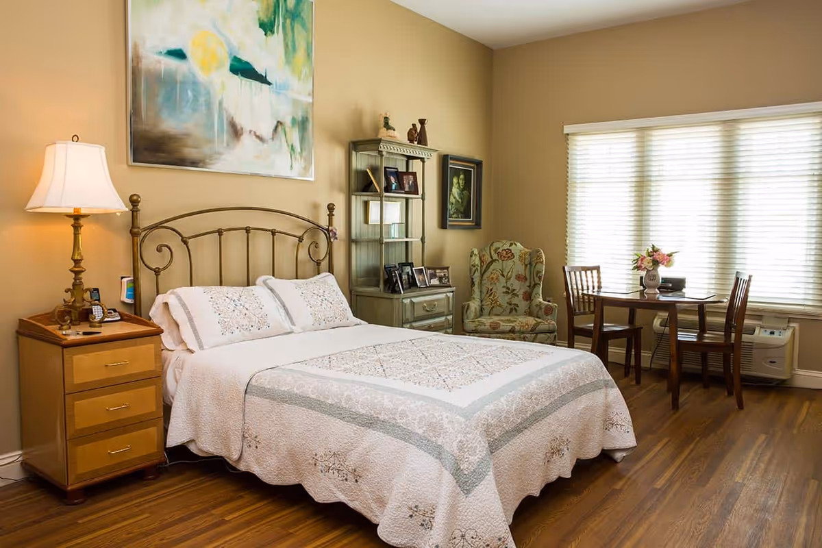 Well-lit furnished bedroom with a bed, nightstand, armchair, small dining table and a large window with blinds.