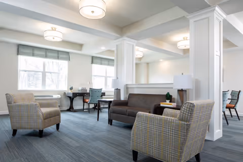 A bright and spacious living room area with large windows letting in natural light. The room features two patterned armchairs, a dark brown sofa, two side tables with lamps, and a desk with a chair near the windows. The walls are painted white, and the ceiling has modern light fixtures. The floor is covered with a blue carpet.