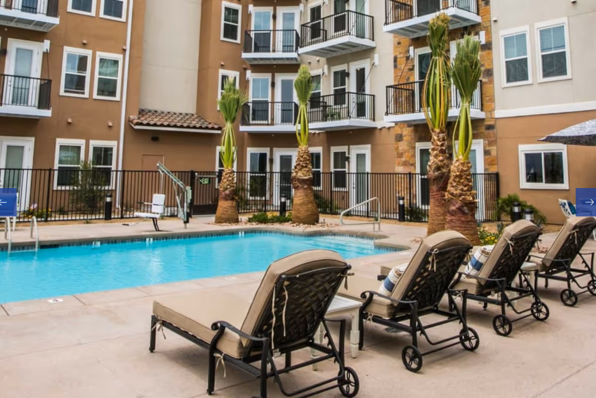Outdoor swimming pool area with lounge chairs, palm trees, and a multi-story residential building with balconies in the background.