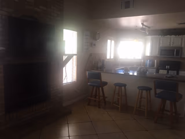 Open-plan kitchen and living area with a breakfast bar and four stools, white cabinets, tiled floor, and a brick fireplace.