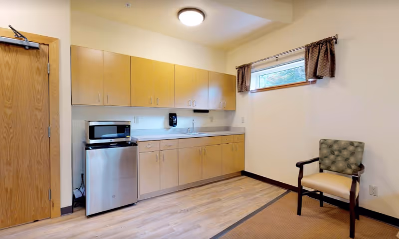 A small kitchenette area with light wood cabinets, a stainless steel mini refrigerator, a microwave, and a sink. There is a single upholstered chair with wooden arms to the right and a small window with brown curtains above it. The floor is a combination of wood and carpet, and the walls are painted light beige.