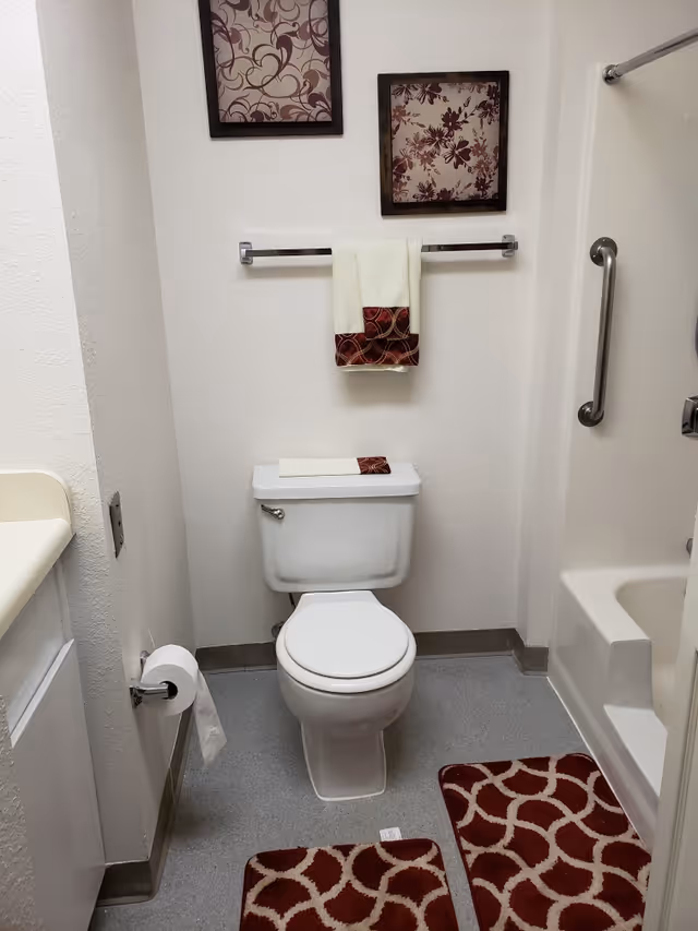 A clean bathroom with a white toilet, a towel rack holding a white towel with a red patterned border, two framed floral artworks on the wall above the towel rack, a bathtub with a grab bar, and two red and white patterned bath mats on the floor.