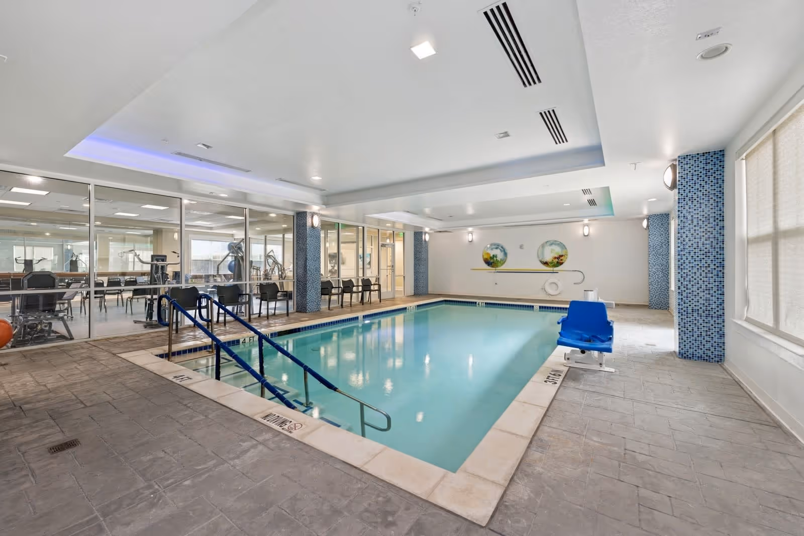 Indoor swimming pool area with a blue pool lift chair on the right side and metal handrails leading into the pool. The pool is surrounded by tiled flooring and has a glass wall on the left side showing a fitness room with exercise equipment and chairs. The ceiling has recessed lighting and a section with blue accent lighting. The walls have blue mosaic tile columns and two round decorative wall pieces at the far end.