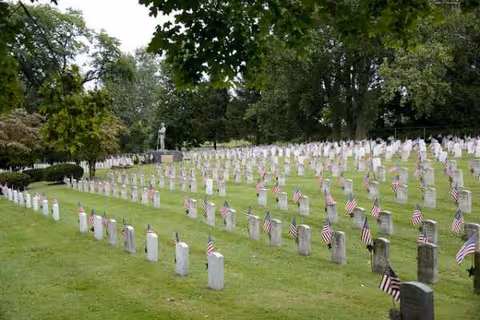 Rows of white grave markers with small American flags on a grassy hillside.