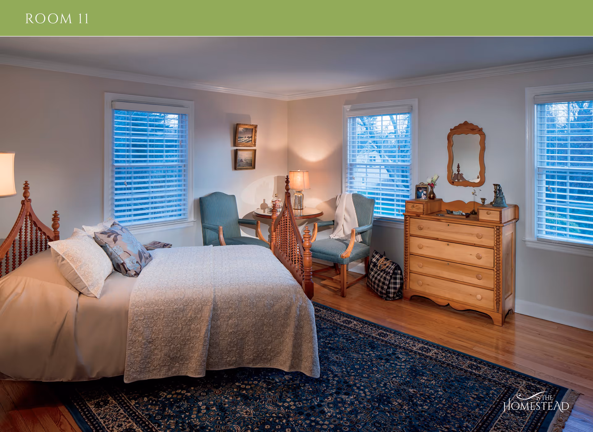 A cozy bedroom with a wooden bed frame, beige bedding, and decorative pillows. The room has three windows with white blinds, two green upholstered chairs around a small round table with a lamp, and a wooden dresser with a mirror above it. A patterned blue rug covers part of the wooden floor. The room is softly lit and has a calm, inviting atmosphere.