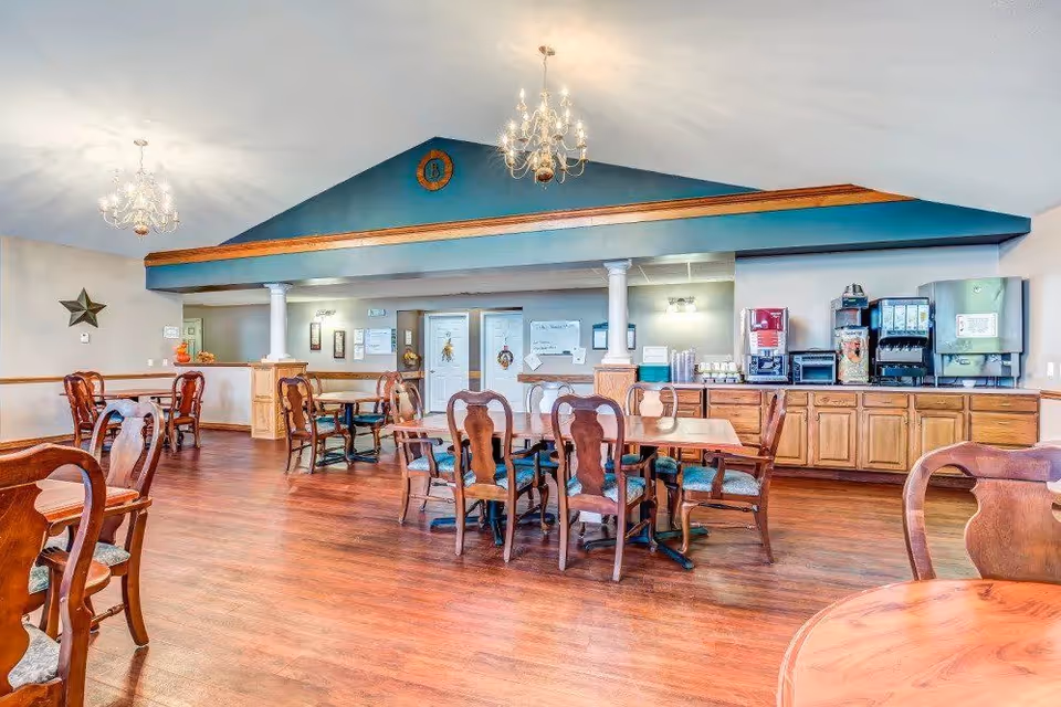 A spacious dining area with wooden floors and multiple wooden tables and chairs. The room features two chandeliers hanging from the ceiling and a blue accent wall with a decorative emblem. Along one wall, there is a counter with beverage dispensers and cups. The room has a warm and inviting atmosphere.
