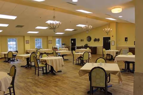 Bright senior dining room with round tables, chairs, and chandeliers in a pale yellow hall.
