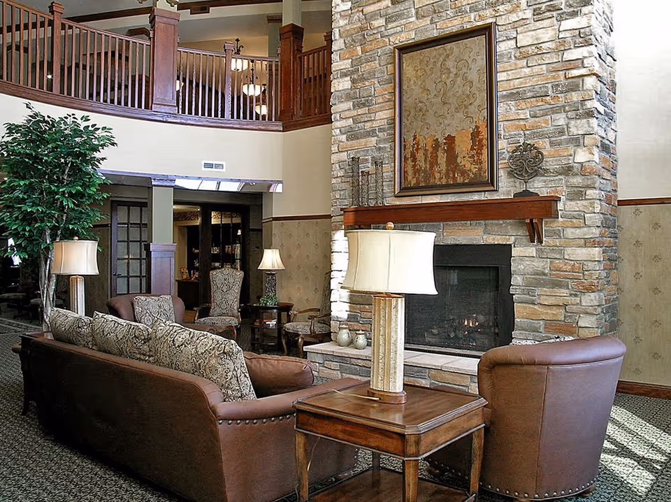 A cozy living room area with a stone fireplace, a wooden mantel holding decorative items, and a framed artwork above it. The room features a brown leather armchair, a patterned sofa with cushions, wooden side tables with lamps, and a tall green plant. The space has a high ceiling with a wooden balcony railing visible above.