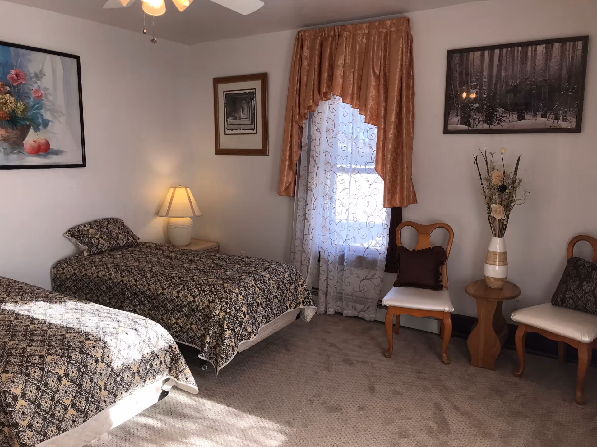 A bedroom with two twin beds covered in matching patterned bedspreads. Between the beds is a small nightstand with a lamp. The room has beige carpet and white walls adorned with three framed pictures. A window with sheer white curtains and a decorative brown valance is on the far wall. Two wooden chairs with cushions and a small wooden table holding a vase with flowers are positioned near the window.