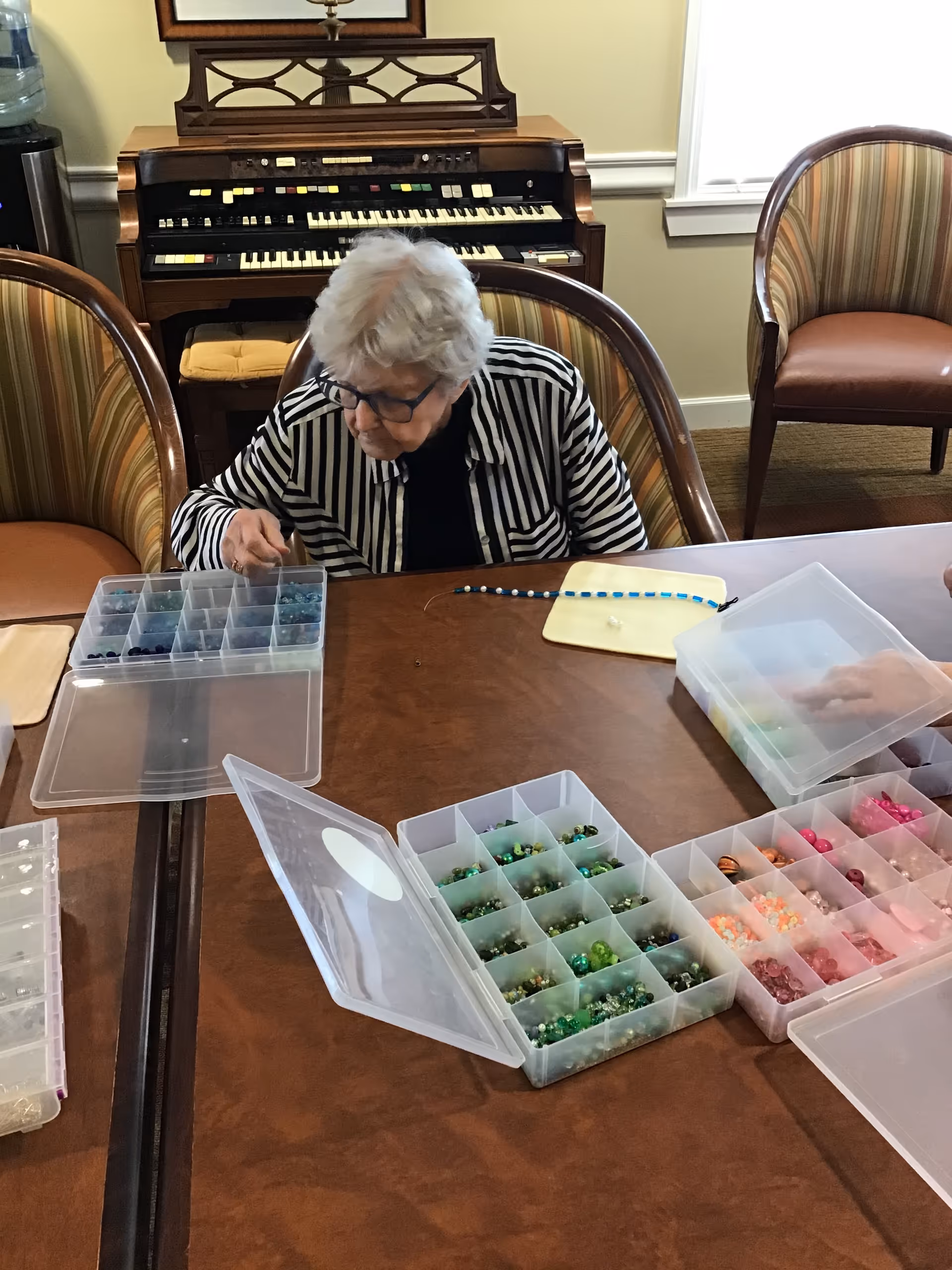 An elderly woman with white hair and glasses is sitting at a wooden table, working on a beading craft project. Several open plastic containers filled with colorful beads are spread out on the table. Behind her is a vintage organ and striped upholstered chairs around the table.