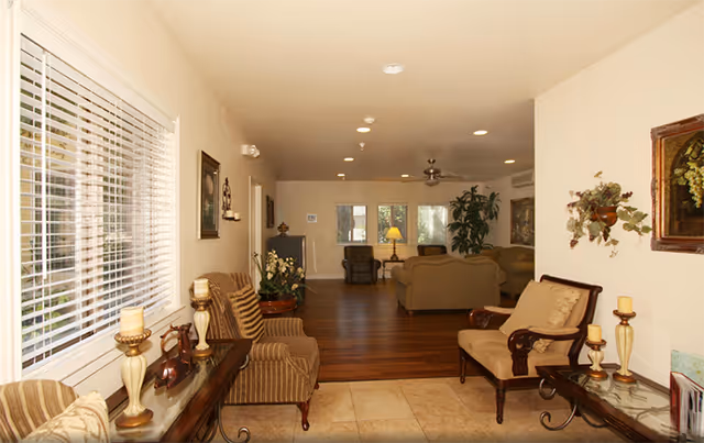 Interior view of a spacious living area with beige walls and a combination of tiled and wooden flooring. The foreground features two cushioned armchairs and a glass-top side table with decorative candles. The background shows a seating area with sofas, armchairs, a lamp, and large windows letting in natural light. There are framed pictures and plants decorating the walls and corners.